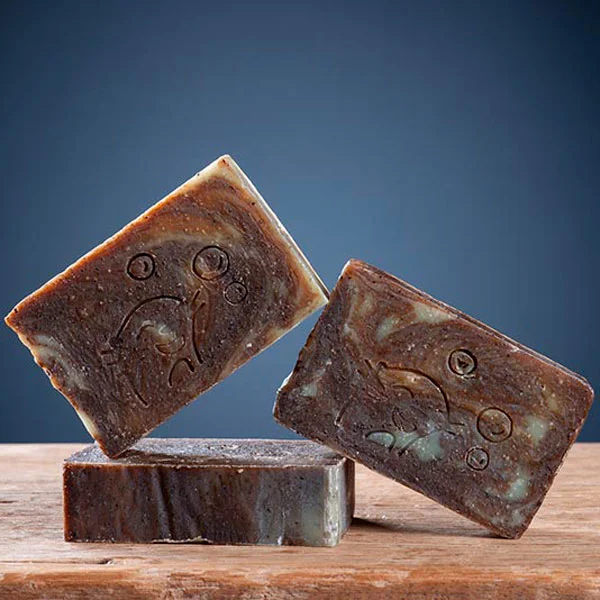 Trois pains de savon café curcuma exfoliant Savonnerie des Monédières en équilibre sur bois, texture granuleuse brun foncé avec marbrage vert argile