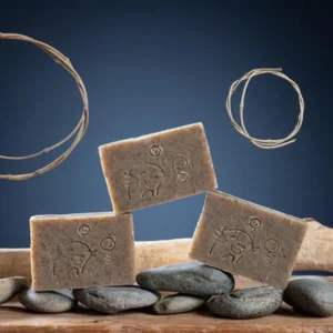 Trois pains de savon olive vanille artisanal Savonnerie des Monédières empilés sur galets, texture naturelle beige-brun avec motif gravé et tiges végétales