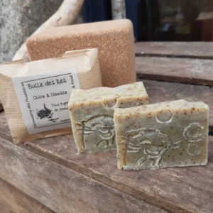 Savon olive vanille Bulle des Îles Savonnerie des Monédières, emballage kraft artisanal, deux pains texturés posés sur bois avec éponge naturelle