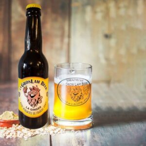 Une bouteille de bière blonde artisanale "La Gros Lam Beer Ginger" avec un logo en forme de houblon souriant, accompagnée d'un verre rempli de bière à la couleur dorée. Sur la table, on distingue des grains de malt et une pincée de gingembre en poudre, évoquant les ingrédients naturels utilisés dans sa fabrication.