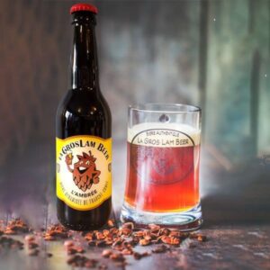 Une bouteille de bière ambrée artisanale 33cl "La Gros Lam Beer" de Brasserie de la Quiotte, à côté d'un verre transparent rempli de la même bière révélant sa couleur ambrée profonde, sur un fond flou qui met en valeur la boisson. Des éclats de malt éparpillés sur la table ajoutent une touche rustique à la scène.