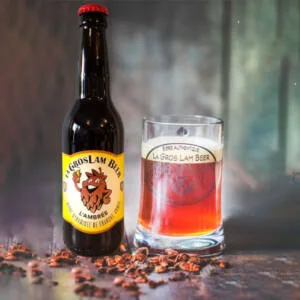 Une bouteille de bière ambrée artisanale 33cl "La Gros Lam Beer" de Brasserie de la Quiotte, à côté d'un verre transparent rempli de la même bière révélant sa couleur ambrée profonde, sur un fond flou qui met en valeur la boisson. Des éclats de malt éparpillés sur la table ajoutent une touche rustique à la scène.