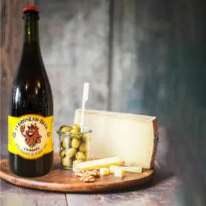 Une bouteille de bière ambrée artisanale de 75 cl "La Gros Lam Beer" de brasserie de la Quiotte présentée sur une planche en bois à côté d'un morceau de fromage à pâte dure, de quelques olives dans un petit pot, et d'un assortiment de noix, créant une composition parfaite pour une dégustation.