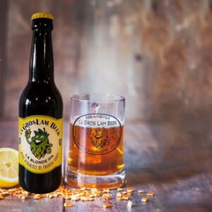 Une bouteille de bière blonde artisanale IPA 33cl "La Gros Lam Beer" avec un logo de houblon, à côté d'un verre rempli de bière. Une tranche de citron et des éclats de houblon parsement la table, évoquant les saveurs agrumes de la bière.