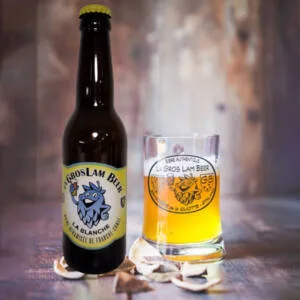 Une bouteille de bière blanche artisanale de 33 cl "La Gros Lam Beer"de brasserie de la Quiotte, à côté d'un verre à moitié rempli de bière présentant une teinte dorée, posés sur un fond rustique en bois. Des écorces d'agrumes sont disposées à côté, suggérant les notes d'agrumes de la bière.