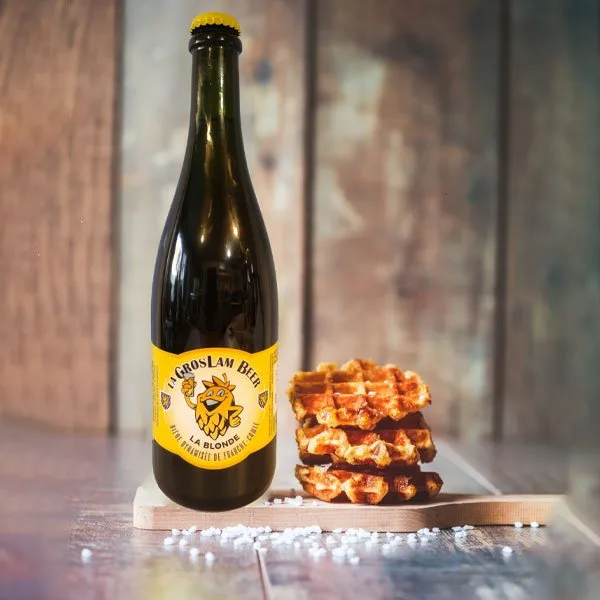 Une bouteille de bière blonde de 75 cl de la Brasserie de la Quiotte à côté d'une gaufre croustillante, posée sur une table rustique en bois. La bière présente une teinte dorée, indiquant sa légèreté et son taux d'alcool. L'étiquette élégante affiche 'Brasserie de la Quiotte', soulignant son origine artisanale. L'association de la bière et de la gaufre évoque une expérience gustative riche et authentique.