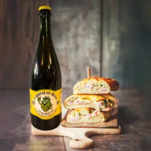 Une grande bouteille de bière blonde artisanale IPA de 75 cl "La Gros Lam Beer" avec un logo de houblon sur l'étiquette est posée à côté d'un sandwich copieux coupé en deux sur une planche en bois, évoquant un accord parfait pour un repas convivial.