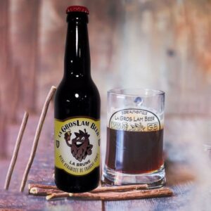 Une bouteille de bière artisanle 33cl brune "Gros Lam Beer" à côté d'un verre rempli de cette bière ambrée, posés sur une surface en bois avec des bâtons de réglisse en arrière-plan. L'étiquette de la bouteille montre un logo ludique le texte "La Brune - Authentique de Franche-Comté"