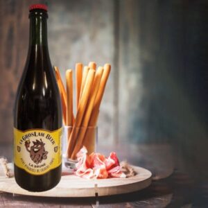 Une grande bouteille de bière brune artisanal 75 cl "Gros Lam Beer" de Brasserie de la Quiotte sur un fond estompé, accompagnée de grissinis dressés dans un verre et de tranches de jambon enroulées sur une planche en bois, évoquant une ambiance de dégustation gourmande.