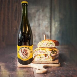 Une grande bouteille de bière blonde artisanale de 75 cl "La Gros Lam Beer Ginger" affiche un logo en forme de houblon souriant sur une étiquette jaune. À côté, un sandwich garni et appétissant est posé sur une planche en bois, suggérant un accord parfait entre la bière et un repas savoureux.