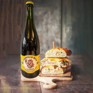 Une grande bouteille de bière blonde artisanale de 75 cl "La Gros Lam Beer Ginger" affiche un logo en forme de houblon souriant sur une étiquette jaune. À côté, un sandwich garni et appétissant est posé sur une planche en bois, suggérant un accord parfait entre la bière et un repas savoureux.