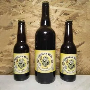 Trois bouteilles de bière artisanale d'Hiver "GrosLam Beer" de tailles 33 cl et 75 cl, avec des étiquettes jaunes montrant un logo de houblon vêtu d'un bonnet d'hiver, alignées sur un fond de bois OSB.