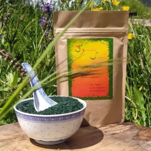 Sachet kraft de spiruline en paillettes croustillantes Spiruline Solaire 100g posés sur une table en bois, avec un bol blanc rempli de paillettes vertes au premier plan, devant les falaises calcaires d'Entrepierres dans les Alpes de Haute Provence