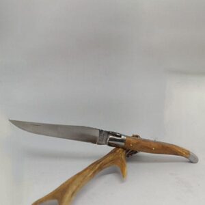 Couteau Laguiole de 12 cm ouvert, montrant une lame en acier inoxydable et un manche en bois d'olivier aux motifs marbrés. La poignée est finement travaillée avec un ressort guilloché visible et un mécanisme de cran d'arrêt. La forme courbée du manche et la finition métallique brillante sur les mitres mettent en valeur le savoir-faire artisanal. Le fond est sobre et uni, concentrant l'attention sur l'artisanat du couteau.