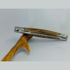 Vue latérale du couteau Laguiole 'L'Outil' de 12 cm fabriqué par Laguiole Laffont, fermé, mettant en évidence le manche en bois de châtaignier aux riches veinures et les mitres en inox satiné. Le couteau repose sur une branche courbée, soulignant la forme ergonomique et la finition soignée de son manche. La photographie capture la texture du bois et le contraste avec l'inox brillant, illustrant la fusion du naturel et du moderne caractéristique de Laguiole Laffont.