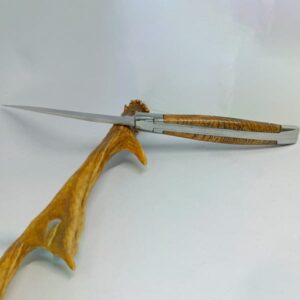 Couteau Laguiole 'L'Outil' de 12 cm ouvert, fabriqué par Laguiole Laffont, posé sur un support en bois naturel. La photo montre le couteau d'un angle mettant en avant la lame tranchante en acier inoxydable et le manche en bois de châtaignier aux détails veinés. Les mitres en inox satiné à l'avant et à l'arrière du manche brillent subtilement, contrastant avec la couleur dorée du bois. L'ensemble évoque la qualité artisanale et la finesse du design de Laguiole Laffont.
