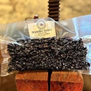 L'image affiche un sachet transparent contenant 500g de myrtilles séchées posé sur un support en bois. En haut du sachet, une étiquette avec un logo circulaire indique "Myrtilles Séchées" et souligne que le produit provient de "Myrtilles du Trimoulet". Les myrtilles déshydratées sont d'une couleur noire intense avec des reflets pourpres, et elles remplissent le sachet de façon à bien montrer leur texture et leur qualité. En arrière-plan, l'environnement évoque un cadre rustique et artisanal.