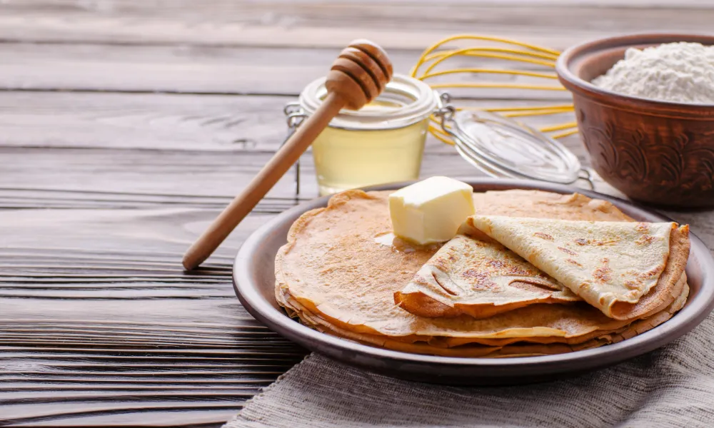 Assiette de crêpes accompagnées de beurre, de miel artisanal et d'ingrédients traditionnels comme la farine, parfaits pour préparer des crêpes à la Chandeleur avec des ingrédients artisanaux.